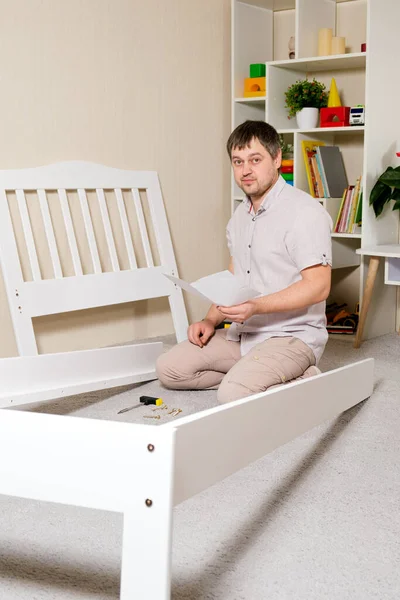 Un hombre con instrucciones para montar muebles va a montar una cuna de madera blanca para niños en un dormitorio para niños. — Foto de Stock