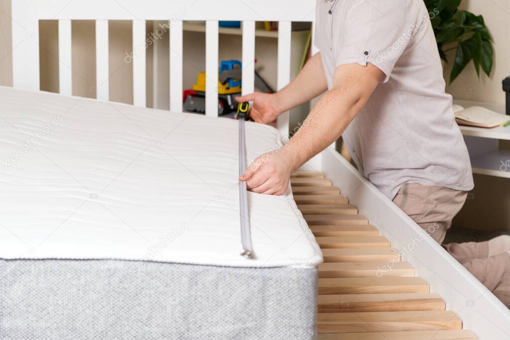 A man measures the length of a childs mattress to mount it on a wooden white bed. Discrepancy between the dimensions of the bed and the mattress.