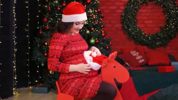 Uma mulher grávida se prepara para o Natal, senta-se em um cavalo de madeira vermelho das crianças e coloca um chapéu em sua barriga. — Vídeo de Stock