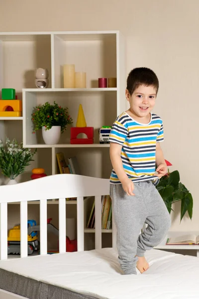 Un niño guapo de 4-5 años salta sobre una cama de madera sobre un colchón, foto vertical —  Fotos de Stock