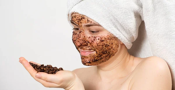Een vrouw met een koffiemasker of scrub op haar gezicht houdt een handvol koffiebonen in haar hand — Stockfoto