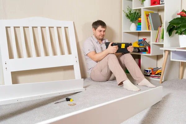 El padre va a armar una cama de madera blanca para niños, se sienta y mira los muebles. — Foto de Stock