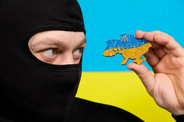 Человек в черной балаклаве держит в руках карту Украины, крупным планом на фоне флага Украины. Концепция войны на Украине, российское нападение — стоковое фото