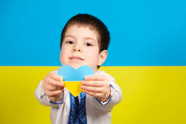 Un niño con una camisa bordada sobre el fondo de la bandera de Ucrania sostiene un corazón amarillo-azul, un lugar para el texto. — Foto de Stock