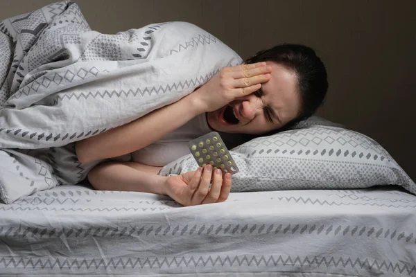 A woman with insomnia lies in bed covered with a blanket, yawns and takes pills for a healthy sleep — Stock Photo, Image