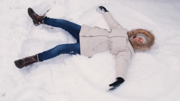 白いジャケットを着た女性が雪の中に横たわって手を上下に上げ、まるで翼を羽ばたき天使を作っているかのように — ストック動画