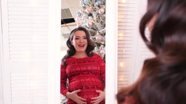 Odbicie w lustrze kobiety w ciąży, oczekująca matka podziwia i głaszcze jej brzuch na tle choinki. — Wideo stockowe