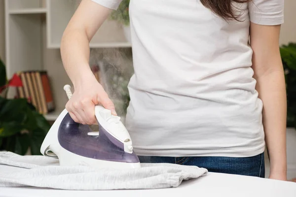 Eine Frau im weißen T-Shirt bügelt Kleidung mit einem Bügeleisen mit einem Dampfer — Stockfoto