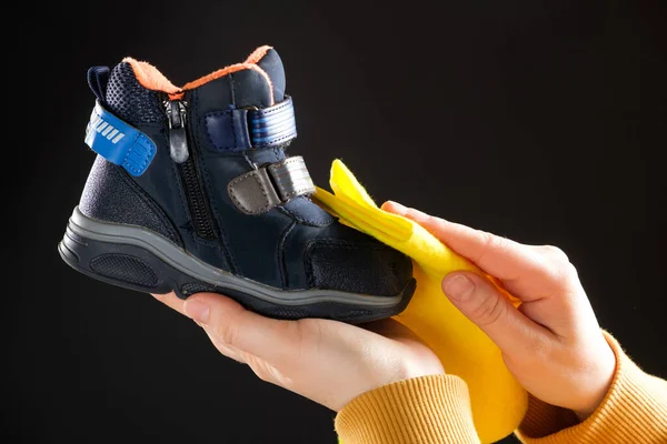 A woman wipes a childs shoe on a black background with a yellow rag. Cleaning from dust and caring for childrens shoes. — Stock Photo, Image