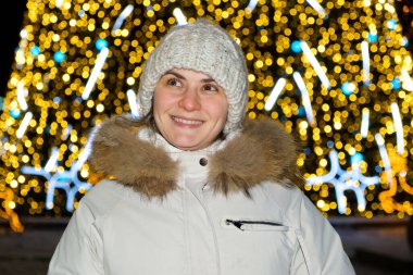 Beyaz şapkalı ve ceketli mutlu bir kadın Noel ağacının yanında duruyor, sarı bokeh, açık havada.