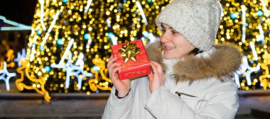 Beyaz kışlık elbiseli bir kadın Noel ağacının önünde duruyor, kırmızı bir hediye kutusu tutuyor ve gülümsüyor..