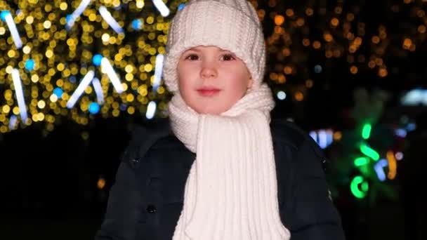 Un hermoso niño preescolar de 4 años se divierte cerca del árbol de Navidad por la noche, mira hacia otro lado y sonríe. — Vídeo de stock