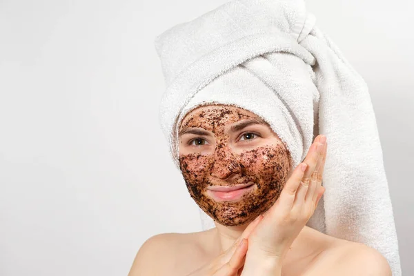 A beautiful young woman with a bath towel on her head and a coffee mask on her face takes care of her skin — Stock Photo, Image