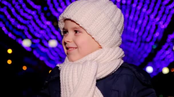 白い帽子のハンサムな子供は夜にクリスマスのガーランドの下に立って笑っています — ストック動画