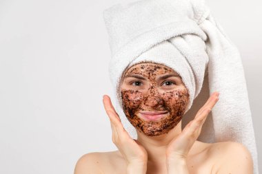 Yüzünde kahve maskesi ve kafasında banyo havlusu olan genç bir kadın gülümsüyor ve beyaz arka plandaki kameraya bakıyor, mesaj atmak için bir yer..