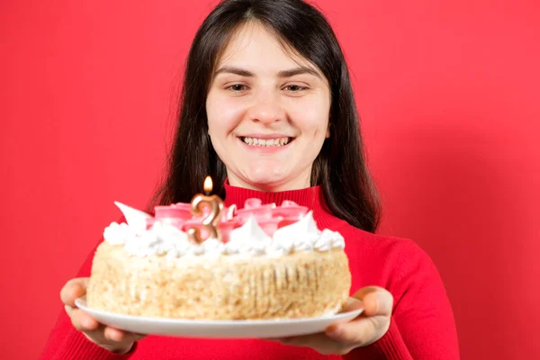 Una mujer feliz sostiene un pastel con una vela en la forma del número tres sobre un fondo rojo. —  Fotos de Stock