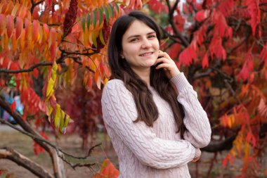 25 yaşında esmer bir kadın uzun saçlarını düzeltir ve sonbaharda kırmızı yaprakların arka planına gülümser.