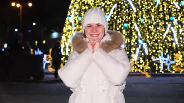A young woman smiles and points her thumbs up near a Christmas tree in the winter in the open air in the evening. — Stockvideo