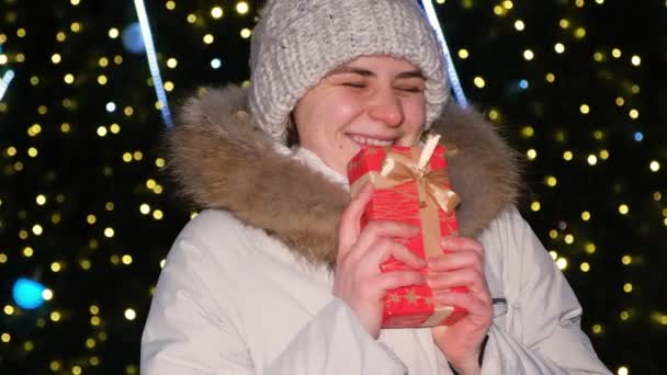 Een jonge vrouw houdt een rode geschenkdoos vast met een strik naast een kerstboom — Stockvideo