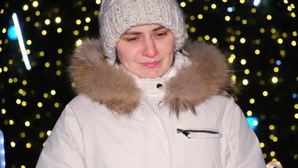 A sad upset woman stands near a Christmas tree with a red gift in her hands — Stockvideo