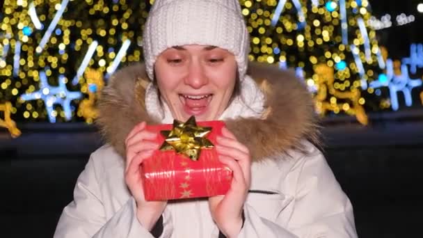 A beautiful woman in a white hat and jacket holds a red gift near the Christmas tree. — Stockvideo