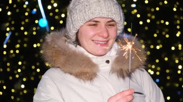 A woman holds a sparkler and smiles standing outside a Christmas tree in the open air — Stockvideo