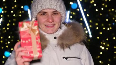 A happy woman shows a red gift box and looks into the camera standing near the Christmas tree