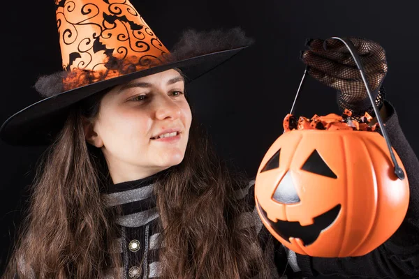 黒い背景の帽子と手袋の魔女の衣装の女性は、キャンディーと笑顔でバケツカボチャを保持しています。ハロウィーンの人々 — ストック写真
