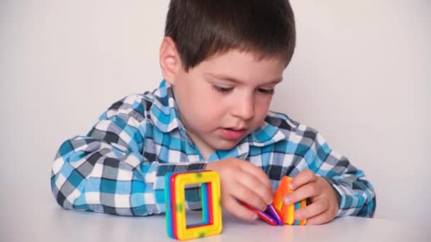 A boy of 4 years old plays with a magnetic constructor, carefully connects the details on a white background — стоковое видео