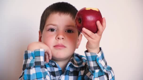 一个4岁的学龄前男孩坐在一个白色背景的桌子旁边，吃着一个大的红色苹果. — 图库视频影像
