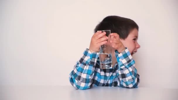 Ένα όμορφο αγόρι 4 ετών πίνει νερό, χαμογελάει και κοιτάζει αλλού, μιλάει, παίρνει ένα ποτήρι στα χέρια του — Αρχείο Βίντεο