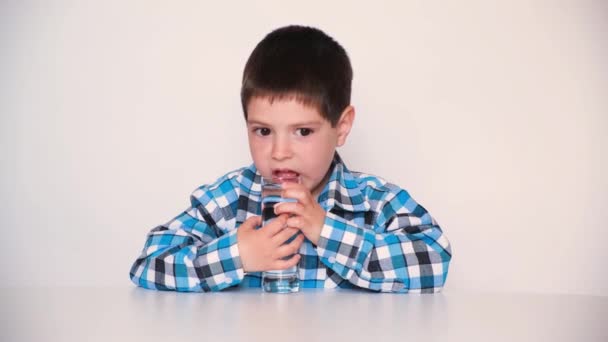 En 4-årig pojke dricker skrattretande vatten ur ett glas, tittar in i kameran och åt sidan, skäller vatten med tungan på en vit bakgrund — Stockvideo