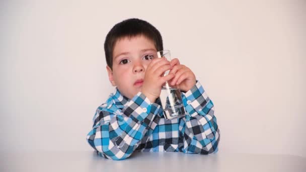 A preschool boy drinks water from a glass, smiles and talks looking into the camera on a white background — стоковое видео