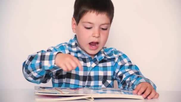 Un niño de 4 años mira un libro con gruesas páginas de cartón con fotos, habla sobre un fondo blanco. — Vídeo de stock