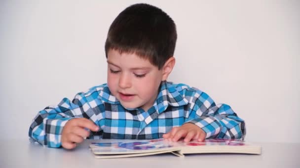 A 4-year-old boy looks at a book with thick cardboard pages with pictures, talks on a white background. — ストック動画