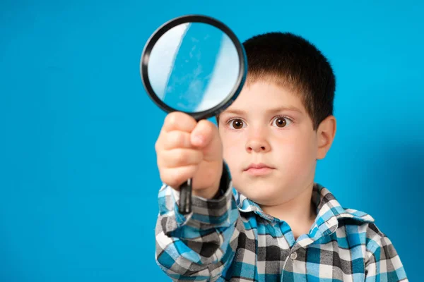 A preschool boy holds in his hand a magnifying glass on a blue background with a place for text. — Stock Photo, Image