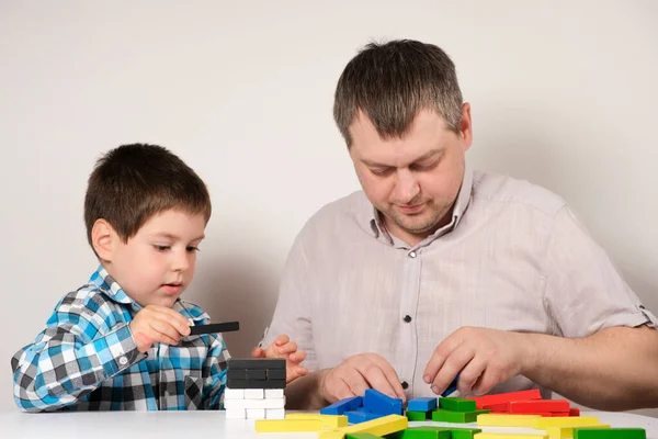 Father and son together build a tower of colorful wooden blocks sitting at a table — Stock Photo, Image