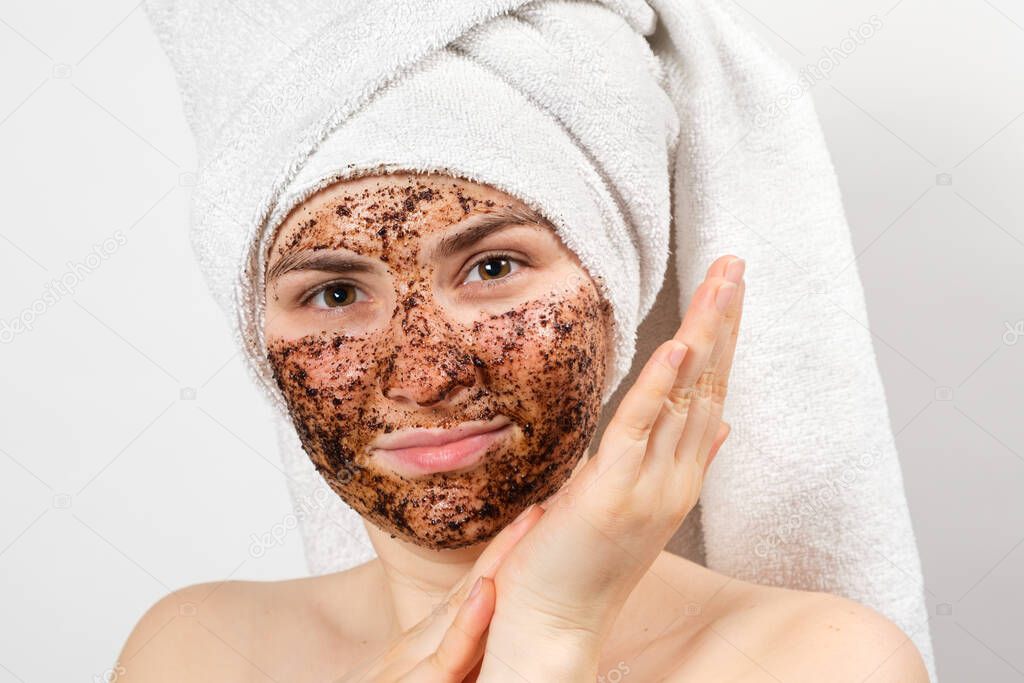 A beautiful young woman with a bath towel on her head and a coffee mask on her face takes care of her skin.