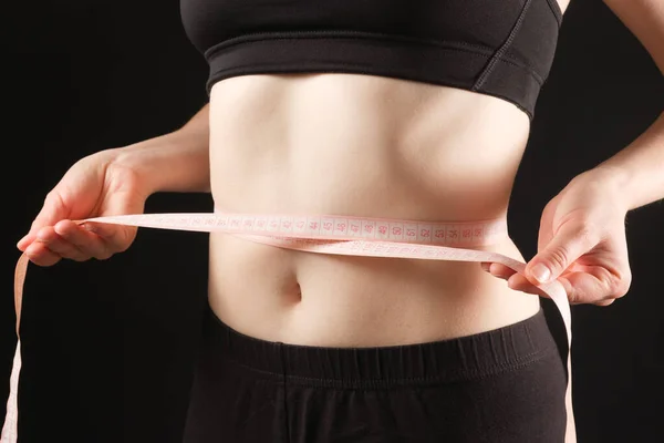 A woman measures the circumference of her abdomen on a black background. The concept of weight loss.