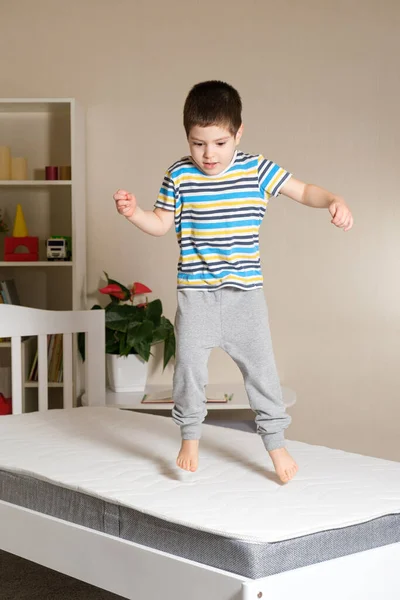 A little handsome boy of 4-5 years jumps on a wooden bed on a mattress, vertical photo.