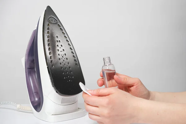 A woman cleans the sole of the iron from soot and scale with a special solution in a bottle and a cotton pad.