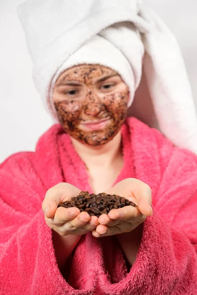 顔にコーヒーマスクをしたピンクのバスローブの女性は手にコーヒー豆を持っています — ストック写真