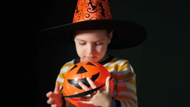 一个戴着万圣节帽子的5岁男孩在一个黑色背景的南瓜形状的桶里窥视着 — 图库视频影像