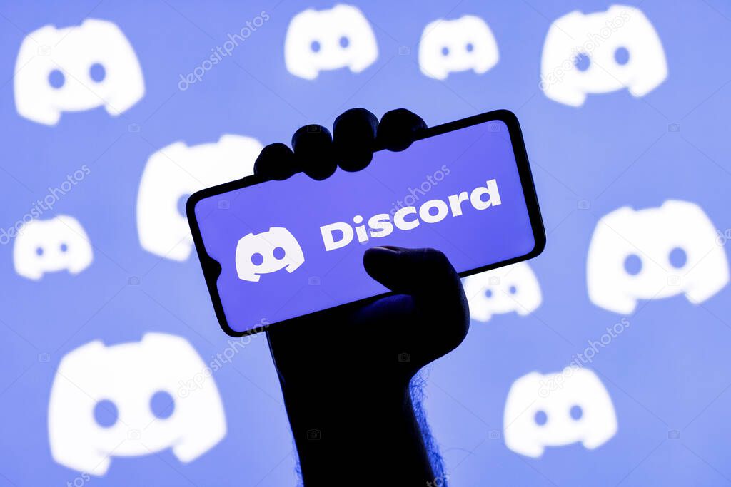 Kazan, Russia - Jan 20, 2022: Smartphone with the Discord social gaming platform logo on the screen in a clenched hand on the background of Discord logos