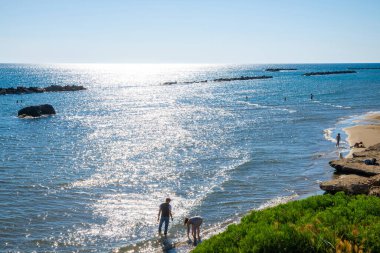 Günbatımından önce banyocularla Anzio plajı. Yazın kayalıklarda mavi gökyüzü ve su üzerindeki ışınlar. Lazio kıyısında, Roma, Lazio, İtalya yakınlarında..