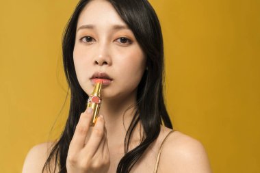 Siyah saçlı kadın altın arkaplanın önüne ruj sürüyor.