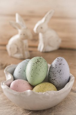 Dekoratif Paskalya yumurtaları pastel renklerde, beyaz el yapımı bir kâse içinde ahşap bir masada, arka planda iki beyaz tahta Paskalya tavşanı.