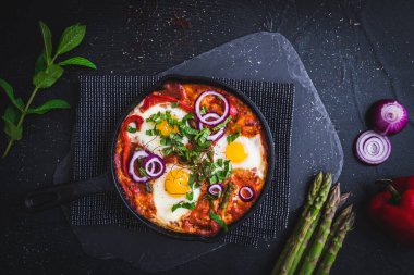 Shakshuka demir bir tavada. Geleneksel Orta Doğu yemeği. Domatesli sahanda yumurta, dolmalık biber, sebze ve otlar, güneşli yumurtalar. Üst görünüm.