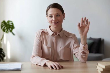 Gülümseyen bayan öğretmen web kamerasına bakıp el sallıyor ve selamlıyor. Çevrimiçi konuşma