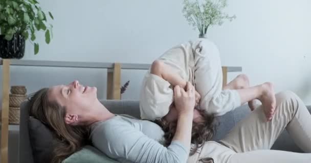 Беззаботный радостный веселый мальчик сын прыгает весело играет и обнимает молодую мать, лежащую на диване — стоковое видео